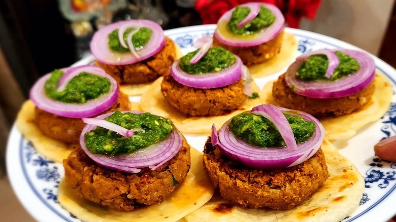 Seven galouti kebabs on plate, each topped with onion and green chutney