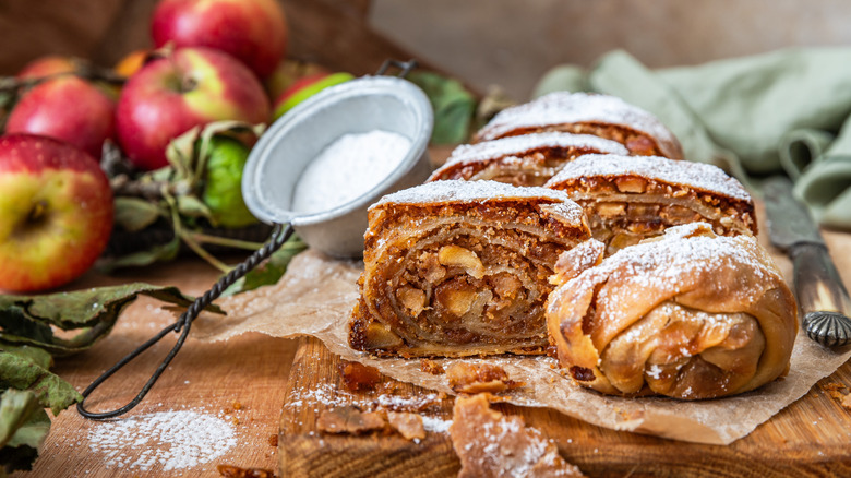 Apple strudel on wooden cutting board surrounded by apples and powdered sugar
