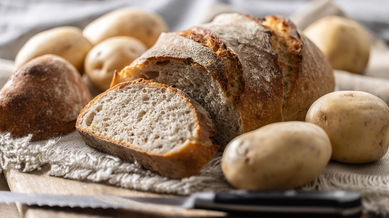 Loaf of bread on cutting board surrounded by potatoes