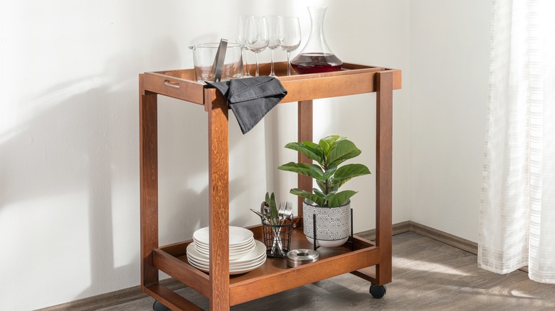 Wooden bar cart with glassware, a decanter with wine, plates, silverware, and a plant.