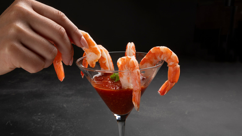 Person picking up a shrimp served with red sauce in a cocktail glass against a dark background