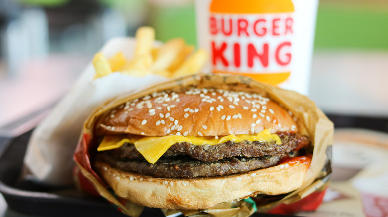 A Burger King double cheeseburger featuring two patties and cheese on a sesame seed bun, wrapped in paper, with fries and a drink out of focus in the background