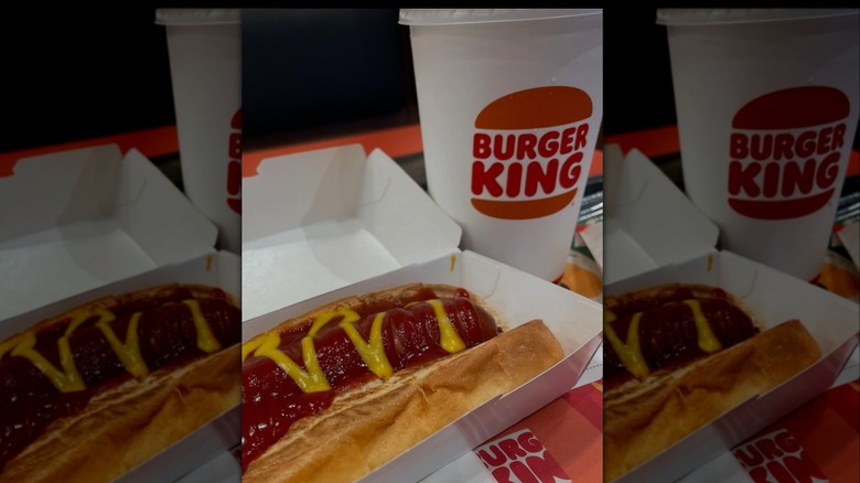 A Burger King hot dog covered in ketchup and mustard, in a cardboard box, with a drink to the side
