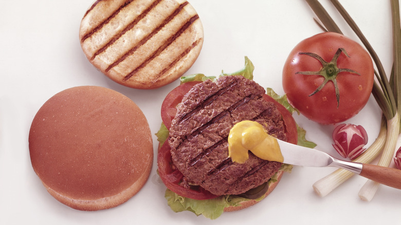 Someone about to spread mustard on a flame broiled  burger with a butter knife. There's a toasted bun, a whole tomato, and green onions arranged around it