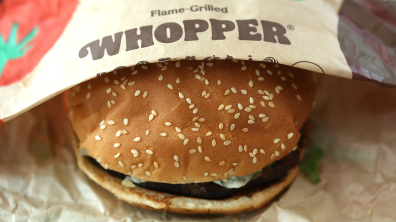 A Burger King Whopper, half covered in paper, showing the sesame seed bun and a bit of the flame broiled patty inside