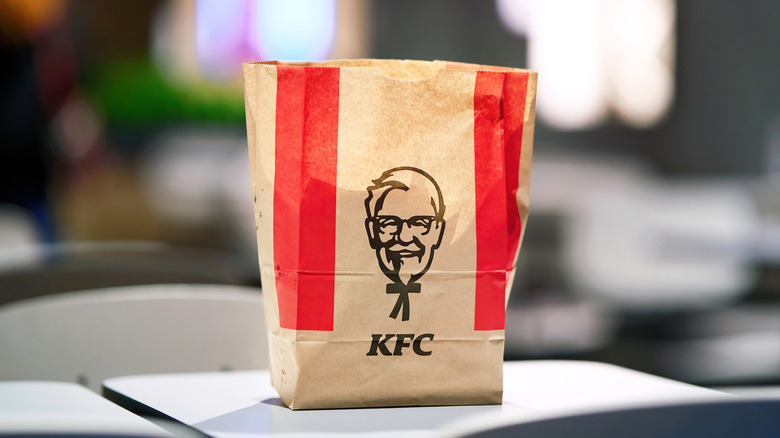 Brown and red paper takeout bag from KFC with Colonel logo sitting on white table