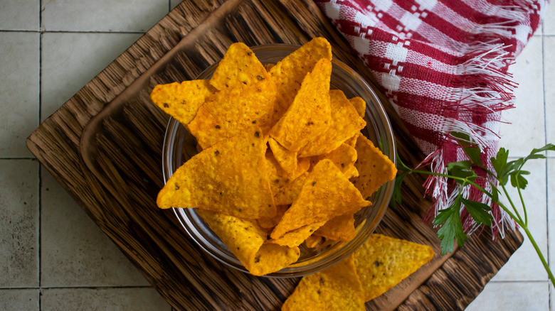 A glass bowl of Doritos rests on a wooden chopping board on a tiled surface next to a kitchen towel