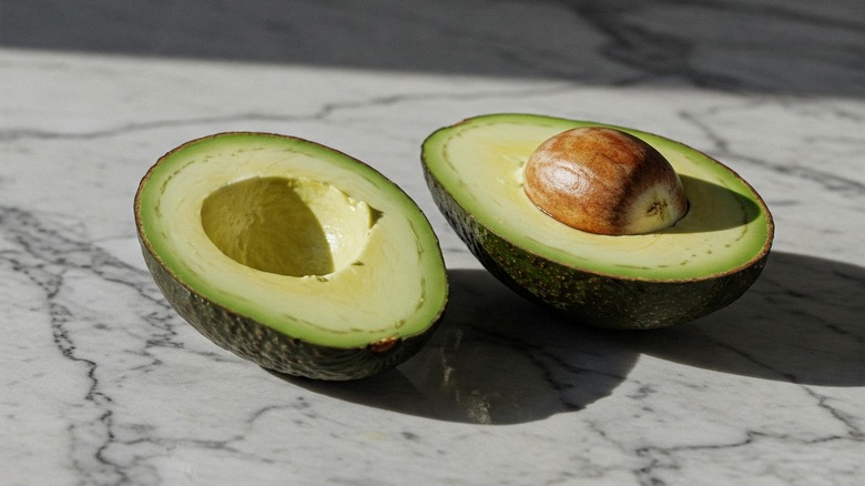 An avocado cut in half on a marble countertop with the pit still attached
