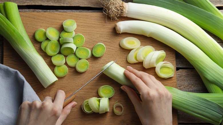 A leek being cut on a cutting board with other leeks around it, including one with its roots intact
