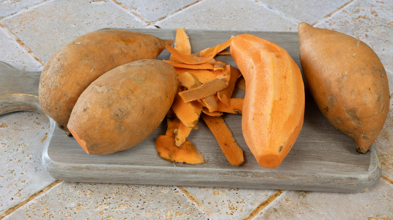 Peeled sweet potato on cutting board, between three unpeeled sweet potatoes