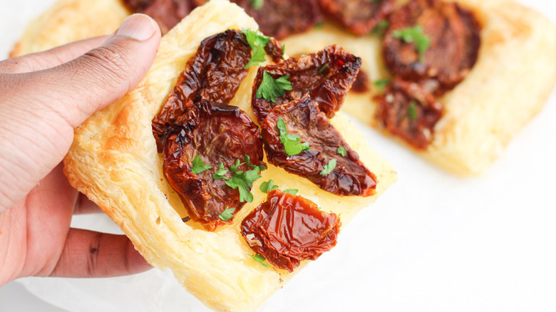 Hand holding slice of sun-dried tomato puff pastry tart