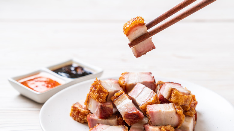 A bowl of pork belly with crispy skin, one piece held by chopsticks