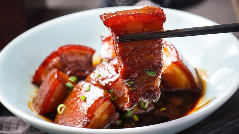 Close-up of glazed pork belly held by chopsticks, bowl with more in background