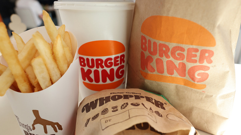 A wrapped Burger King Whopper in front of a Burger King drink, fries, and bag