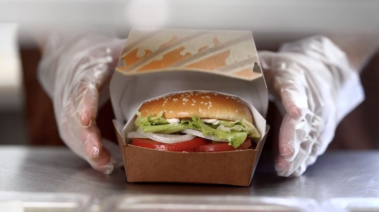 A 2026 Whopper served in a protective clamshell box being presented by someone wearing gloves