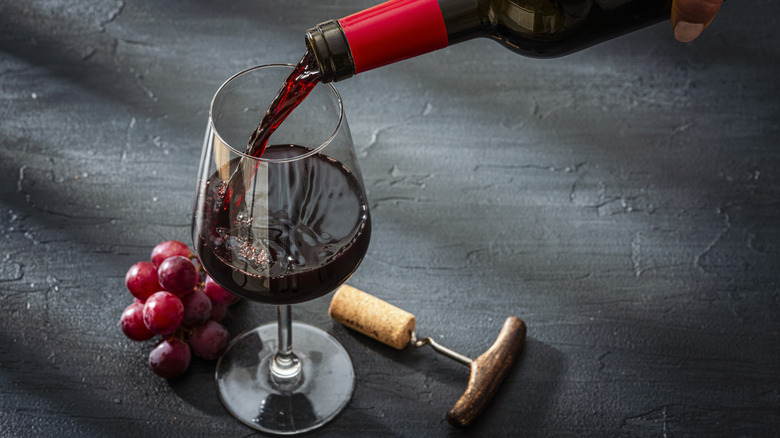 Pouring red wine into a glass with opener and grapes on a dark counter