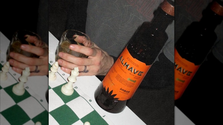 Bottle of Almave Ámbar next to hand holding glass of drink, green and white chessboard beside