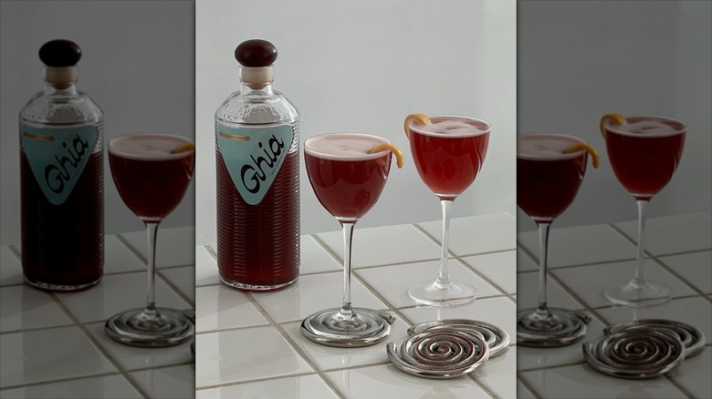 Bottle of Ghia aperitif next to two glasses with drink, orange peel garnish