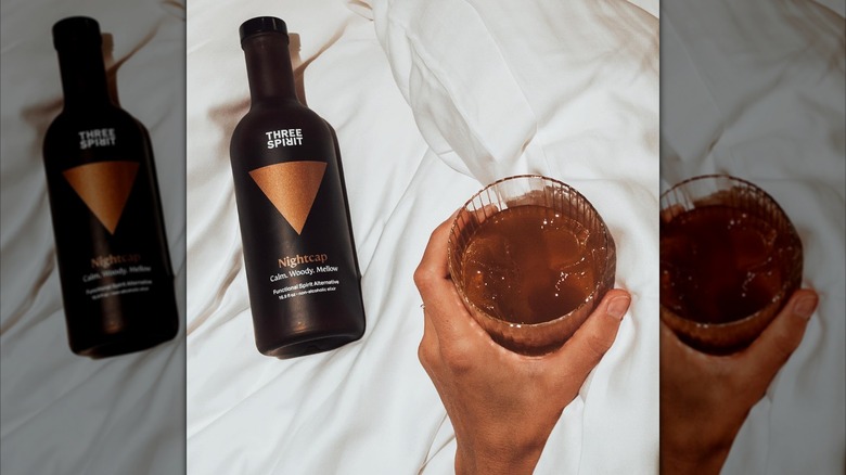 Bottle of Three Spirit Nightcap on white fabric surface, hand holding drink in glass beside