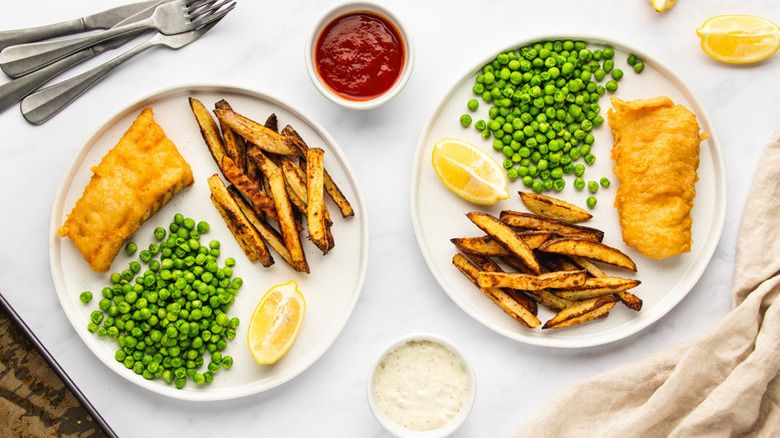 Fish, chips, and peas on two plates next to pots of ketchup and tartar sauce