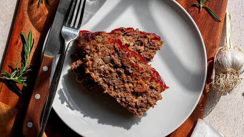 top view of a sliced meatloaf on plate next to utensils, garlic, and rosemary