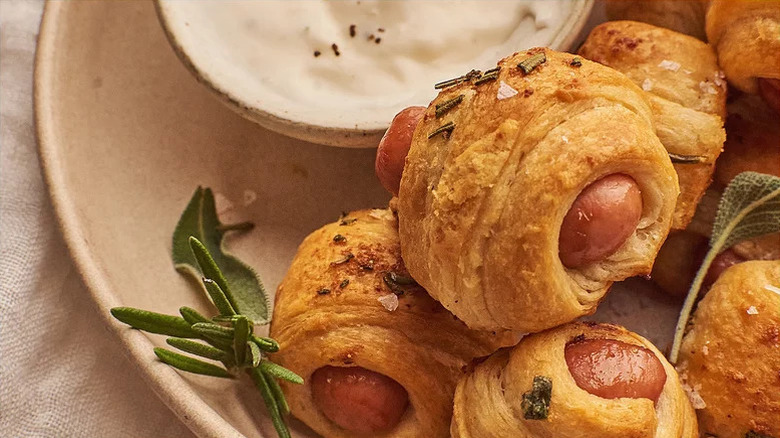 herby pigs in a blanket stacked on a plate with greens and a white dipping sauce served on the side