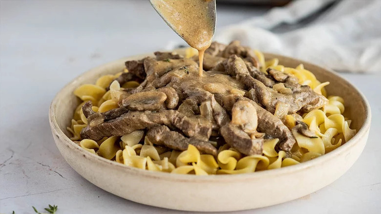 spooning sauce over creamy beef Stroganoff served over pasta in a bowl