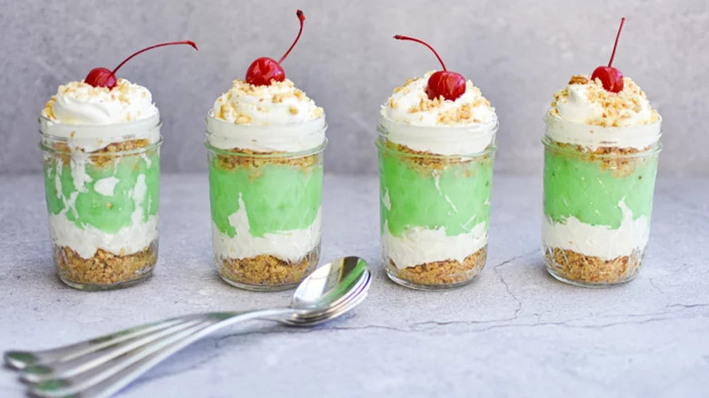 Individual pistachio torte in small glasses with silver spoons topped with cream and maraschino cherries