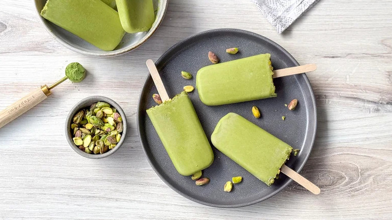 Matcha pistachio popsicles arranged on grey plate with whole pistachios sprinkled around and served in a bowl