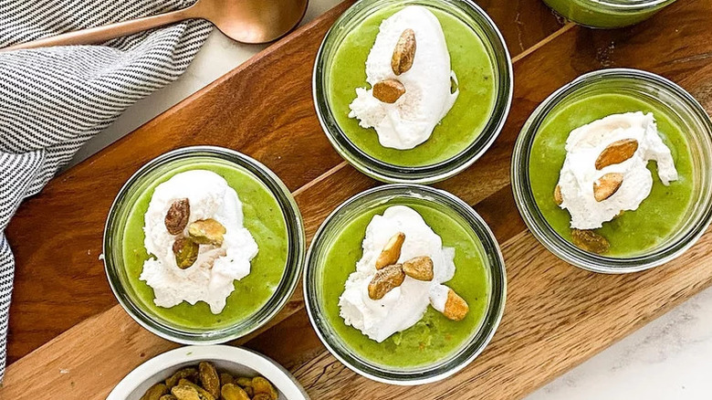 Pistachio pudding in small glass jars on wooden board topped with cream and whole pistachios