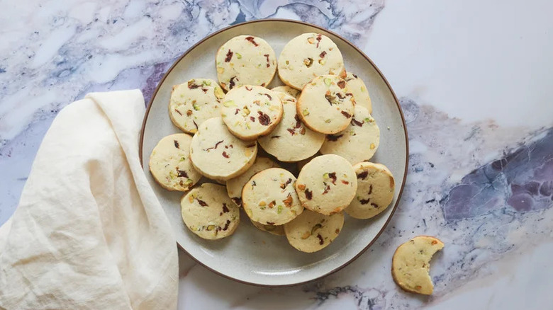 Cranberry pistachio cookies stacked on beige plate with a kitchen towel on a marbled counter