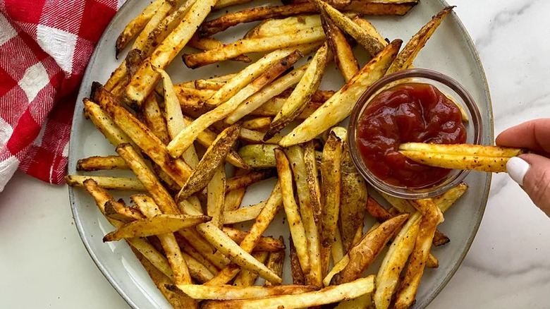 A hand dipping air fryer French fries laid out on a beige plate into a glass jar with ketchup