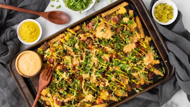 Bacon cheeseburger loaded fries on large dark baking sheet covered in various toppings with more garnishes served in bowls on the side