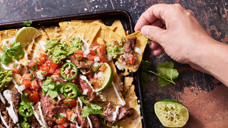 Sheet pan of carne asada nachos with hand removing topping-loaded tortilla chip