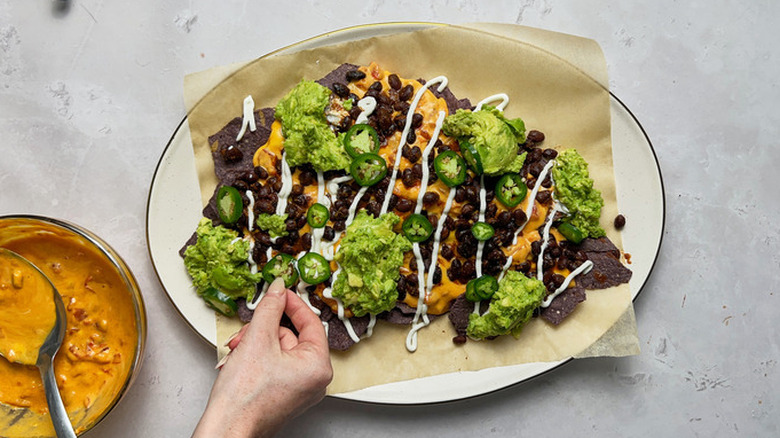 Hand adding jalapeno slices to platter of blue corn tortilla nachos, topped with guacamole and pumpkin queso