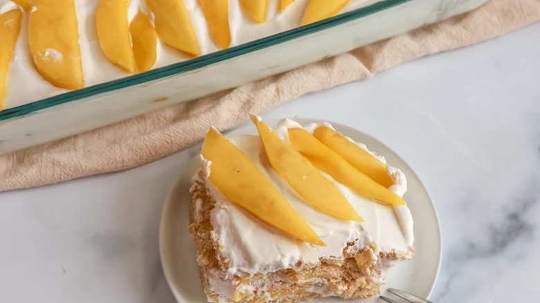 A slice of mango float on a plate next to the rest of the cake in a glass dish