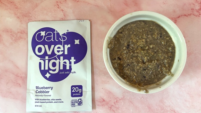 Bowl of Oats Overnight Blueberry Cobbler next to packet on pink surface