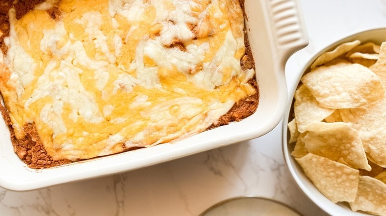 Cheesy bean dip in baking dish next to bowl of tortilla chips