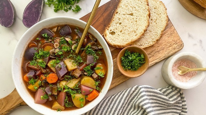 Bowl of mushroom pot roast with purple potatoes and carrots, next to slices of bread and bowl of chopped parsley