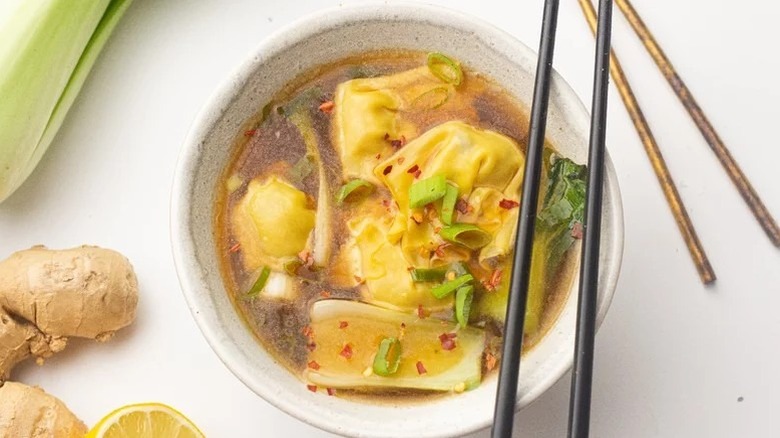 Bowl of wonton soup with chopsticks, surrounded by ginger, lemon, and baby bok choy