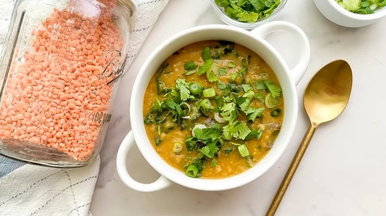 Red lentil soup in bowl, topped with cilantro and green onions, next to jar of red lentils and spoon
