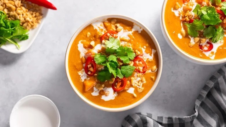 Two bowls of coconut peanut soup topped with peppers, cilantro, and coconut cream