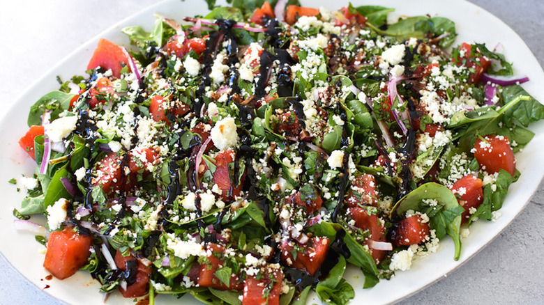 Spinach and watermelon salad on white plate, garnished with crumbled Cotija cheese and balsamic glaze