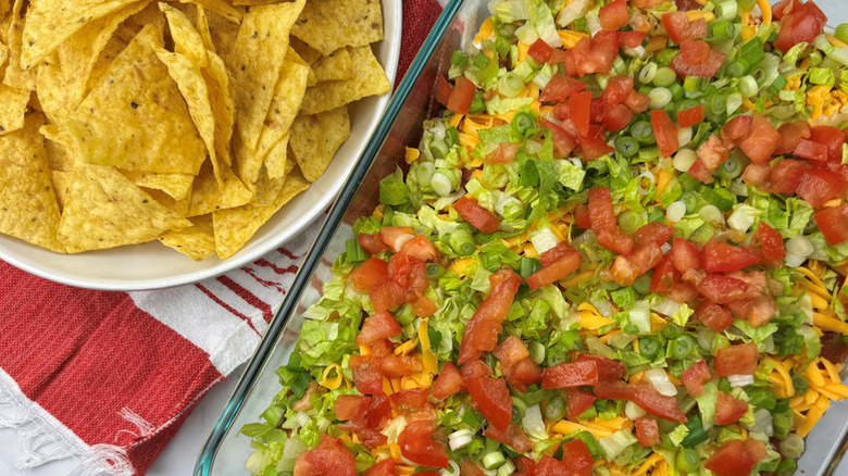 Taco dip in glass casserole dish next to bowl of tortilla chips