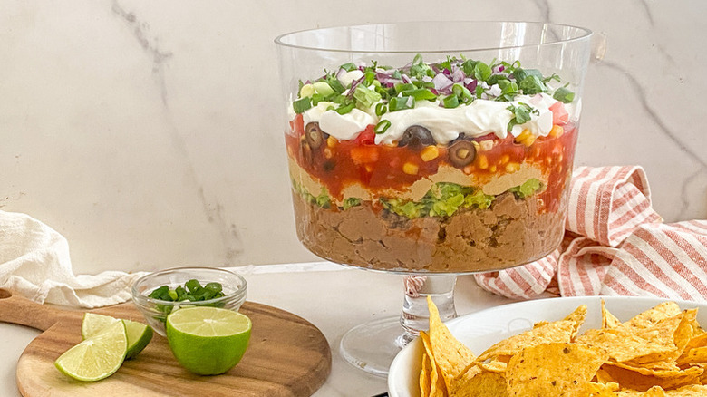 7-layer dip in glass trifle dish, next to bowl of tortilla chips