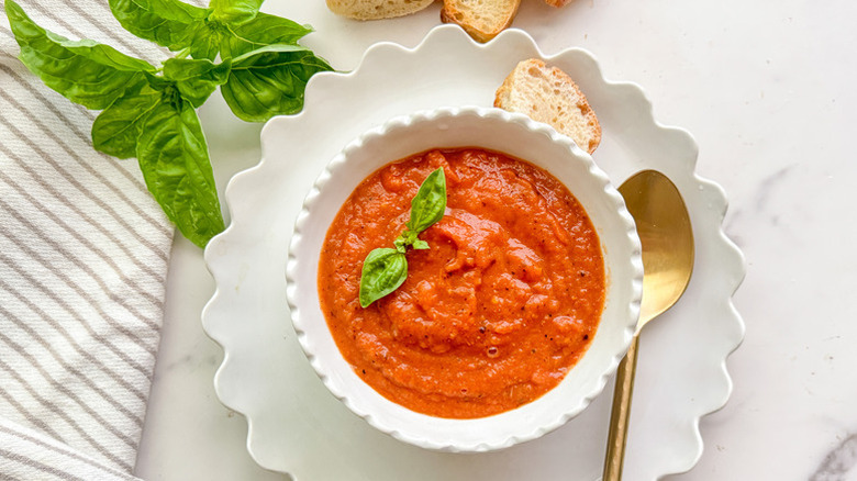 top view of a white bowl filled with tomato soup with bread and fresh basil