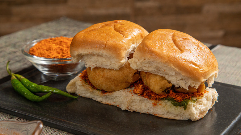 Two Vada Pav sandwiches on black slate with peppers and seasoning