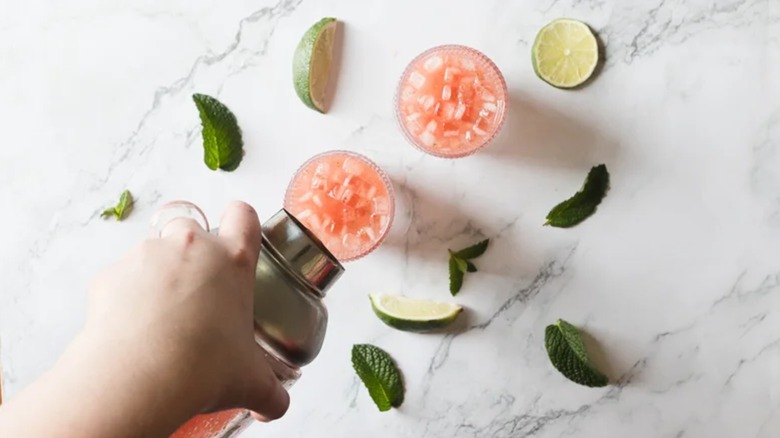 Hand pouring watermelon cocktail into coupe glasses on marble tabletop