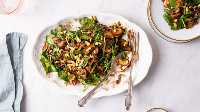 Wilted spinach, mushroom, and pine nut salad on white serving platter with fork and spoon