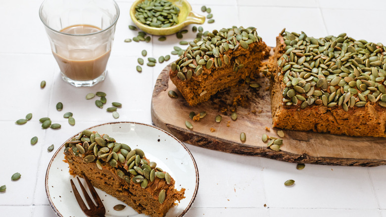 A slice of pumpkin bread on small plate next to sliced pumpkin bread on a cutting board next to cup of coffee